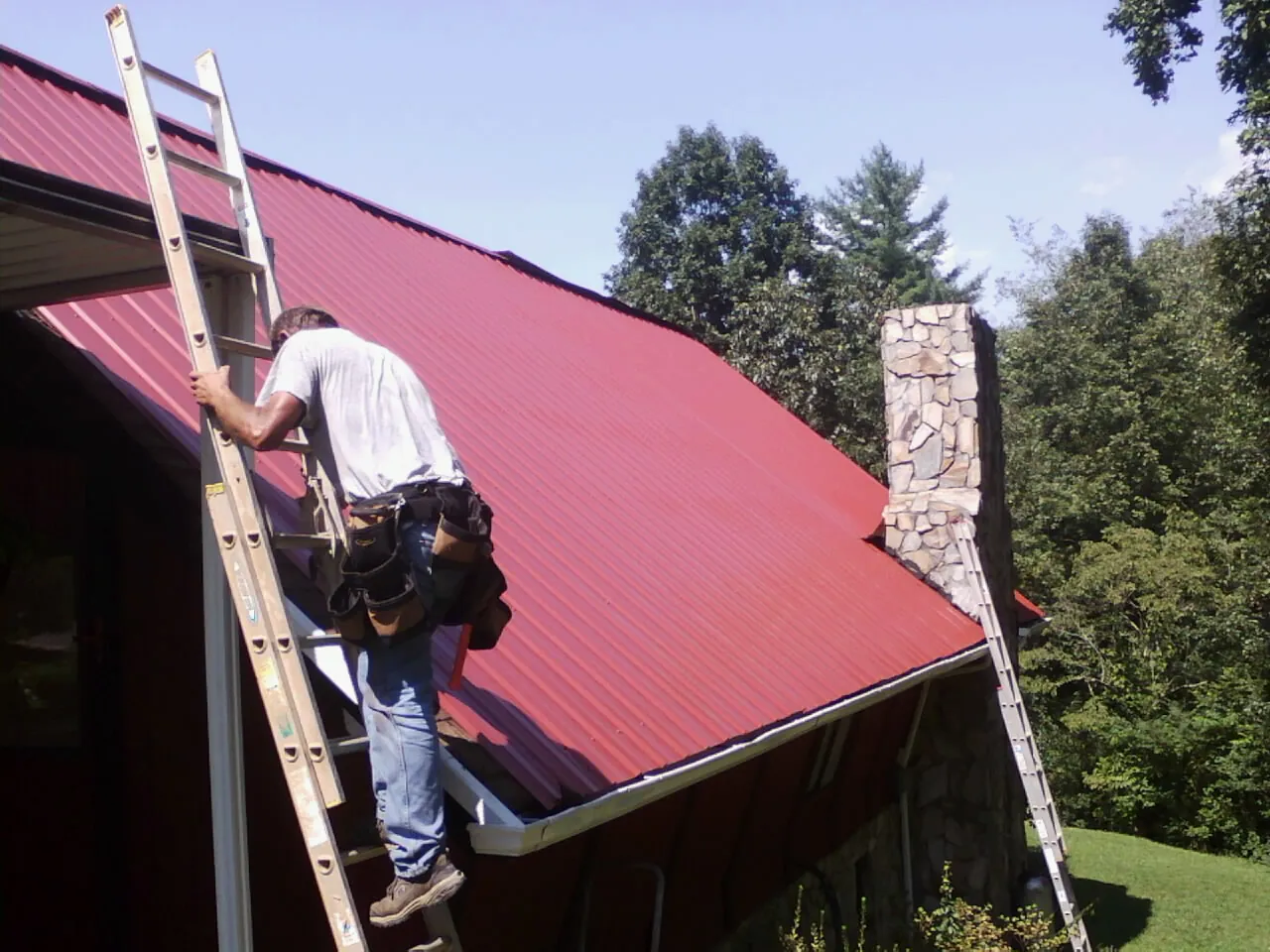 Roofer on a ladder beside a red metal roof and stone chimney — Soffit & Fascia Repair by Roofing Brothers in Delano
